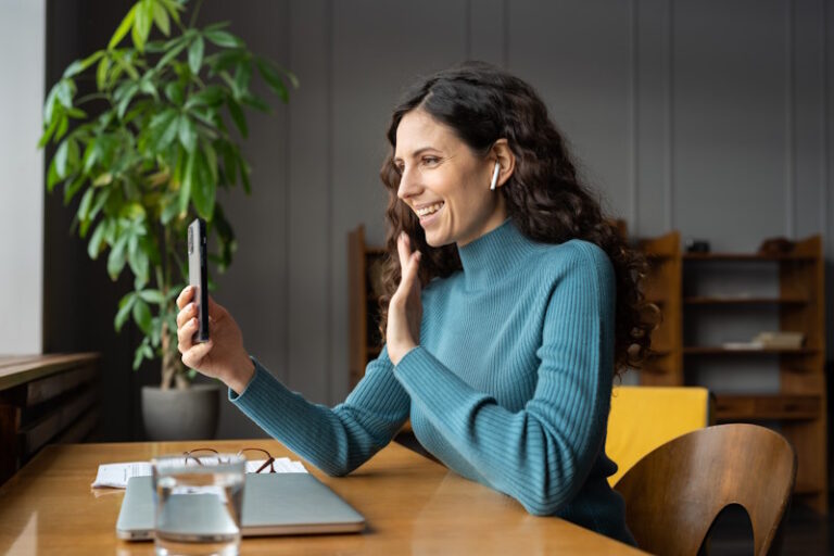 Young happy smiling female office worker enjoying video chat on smartphone with friend of family during work break, positive businesswoman wearing earbuds greeting waving with hand at mobile camera